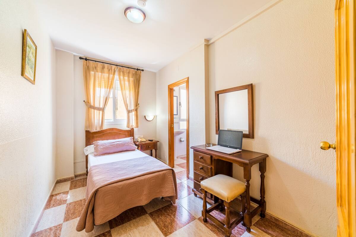 A bedroom with a bed, a desk with a laptop, a chair, and a mirror.