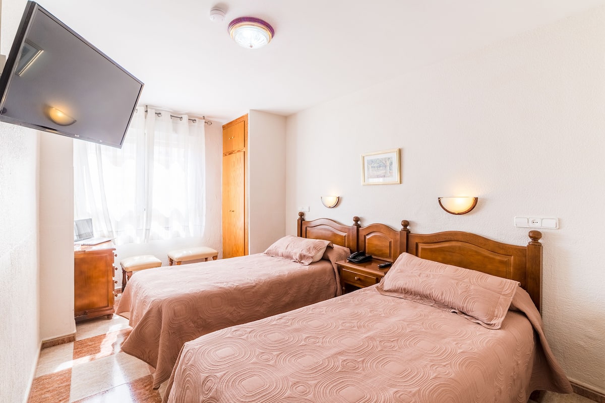 A hotel room with two beds, a TV, a desk, and a window with curtains.
