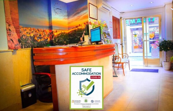 A hotel reception area with a 'Safe Accommodation' sign, a computer monitor, and a pharmacy visible through the glass doors.