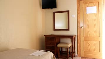 A bedroom with a bed, a wooden desk with a chair, a television mounted on the wall, and a wooden door.