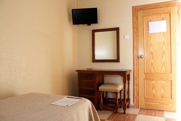 A bedroom with a bed, a wooden desk with a chair, a television mounted on the wall, and a wooden door. - Hotel Sevilla (Almeria)