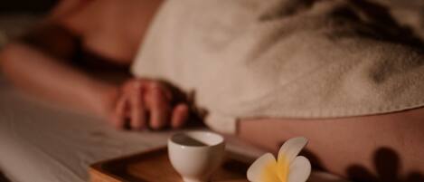 Una persona sdraiata su un letto con un asciugamano, un vassoio con un fiore e una tazza.