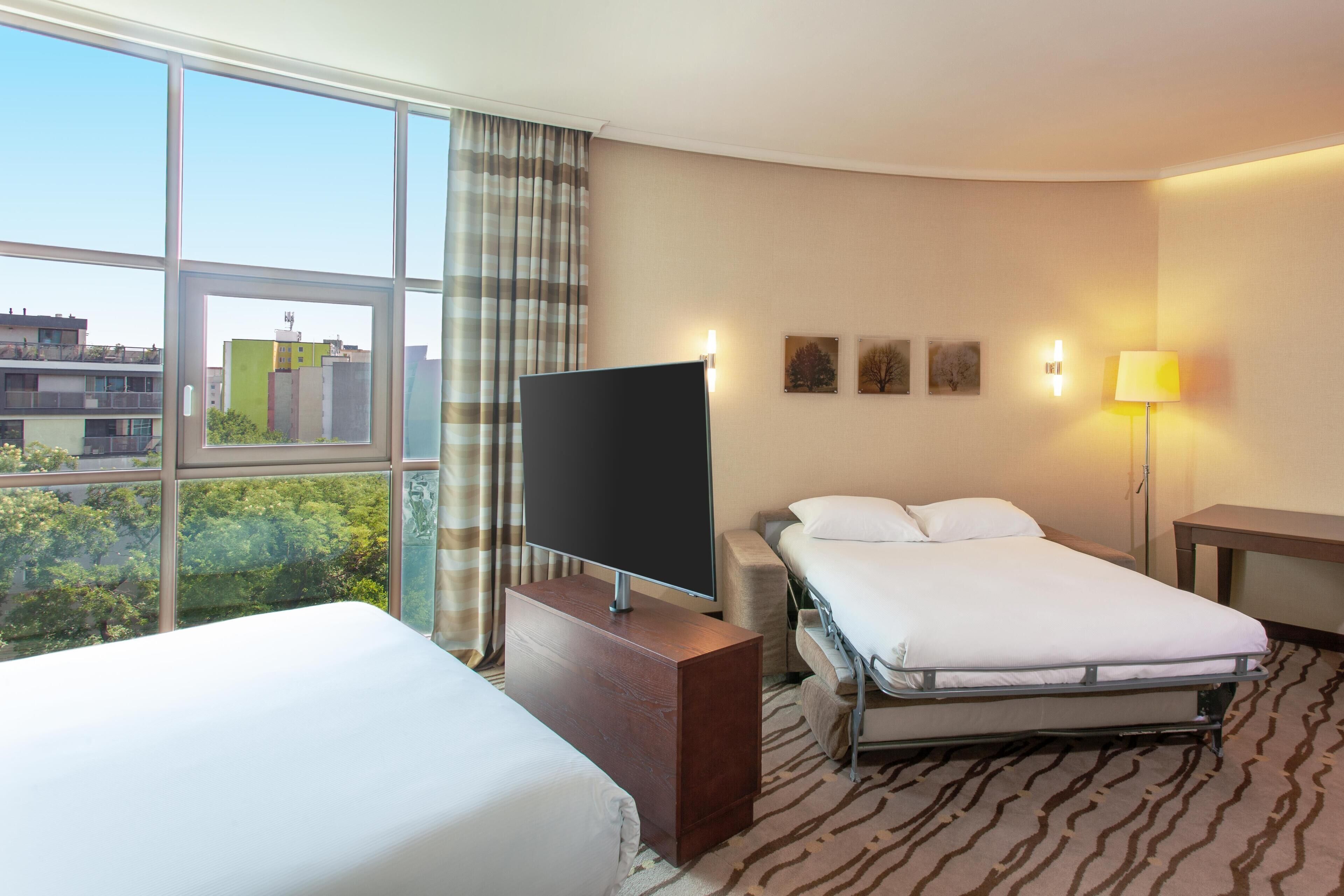 A hotel room with a large window, a flat-screen TV, a bed, a bedside table, and a lamp.