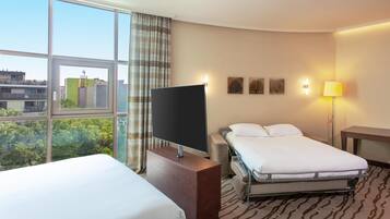 A hotel room with a large window, a flat-screen TV, a bed, a bedside table, and a lamp.