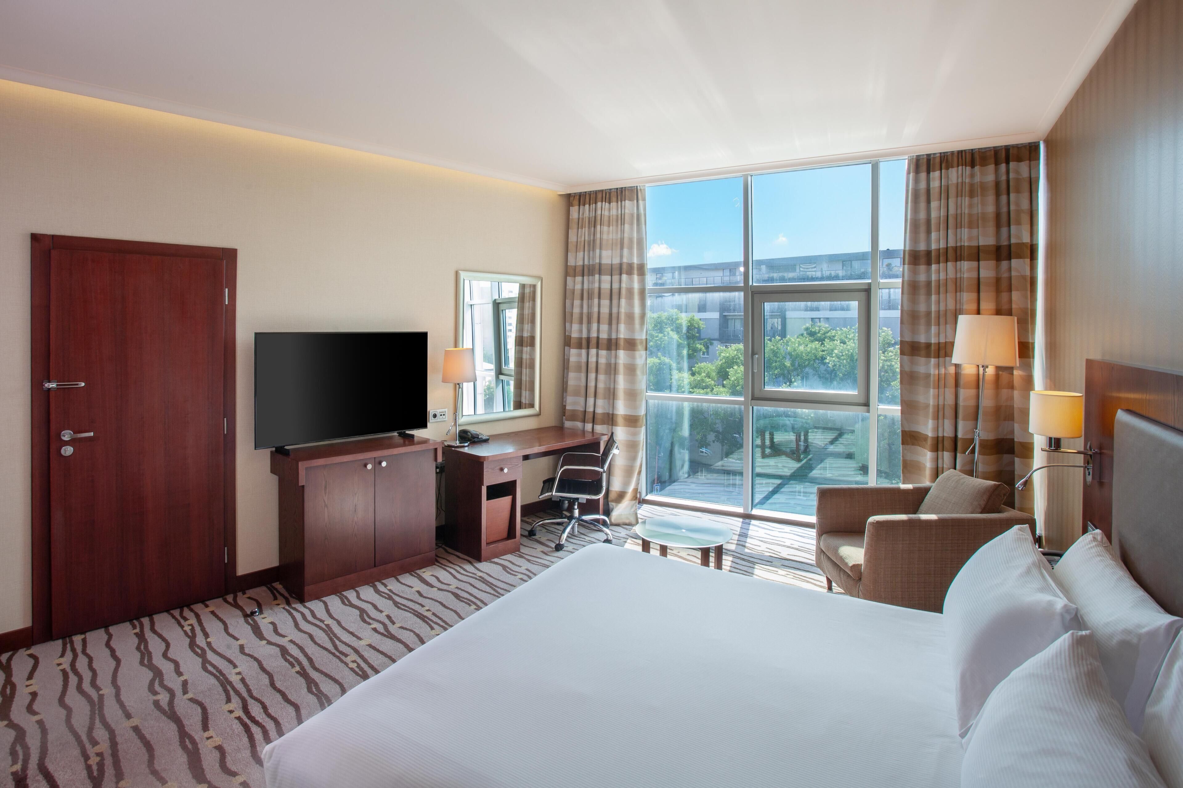 Habitación de hotel con una cama grande, un escritorio con una silla, un televisor y vistas al exterior a través de amplias ventanas.
