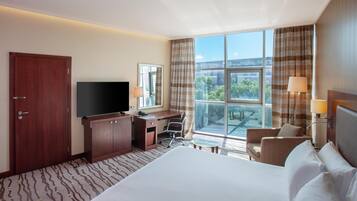 Habitación de hotel con una cama grande, un escritorio con una silla, un televisor y vistas al exterior a través de amplias ventanas.