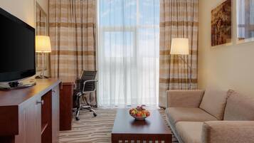 A hotel room with a TV, a desk, a chair, a couch, a coffee table with fruit, and curtains.