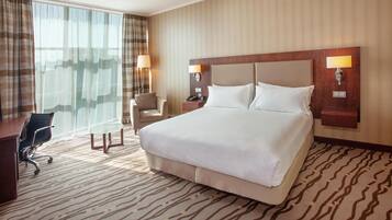 Habitación de hotel con una cama grande, un escritorio con silla, una lámpara y vistas al exterior.