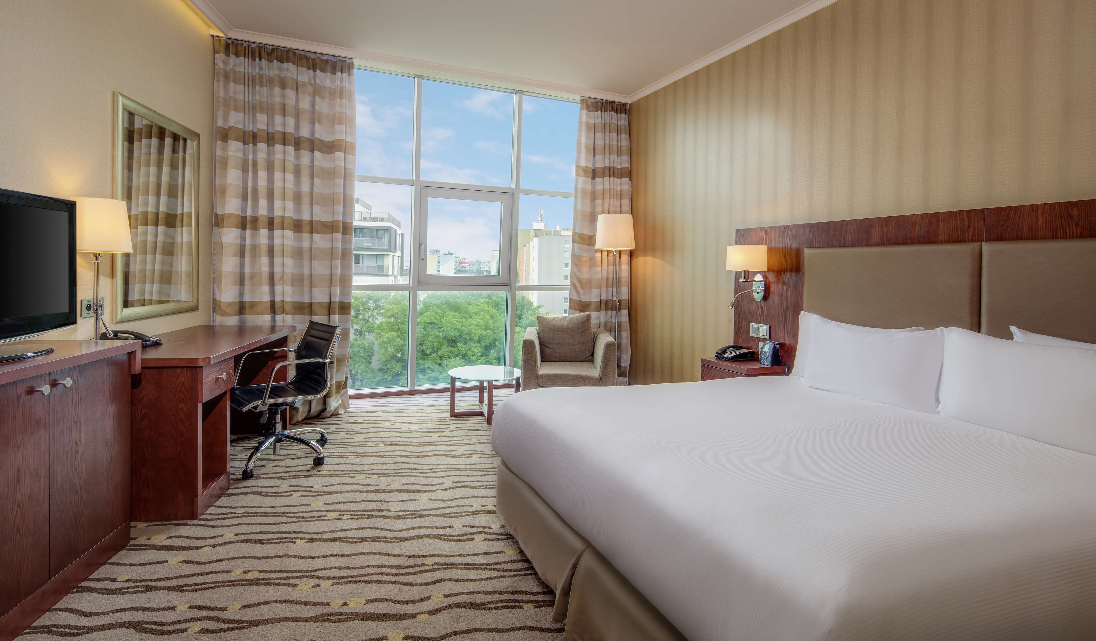 A hotel room with a large bed, a desk with a chair, a TV, a small table, and a nightstand with a lamp.