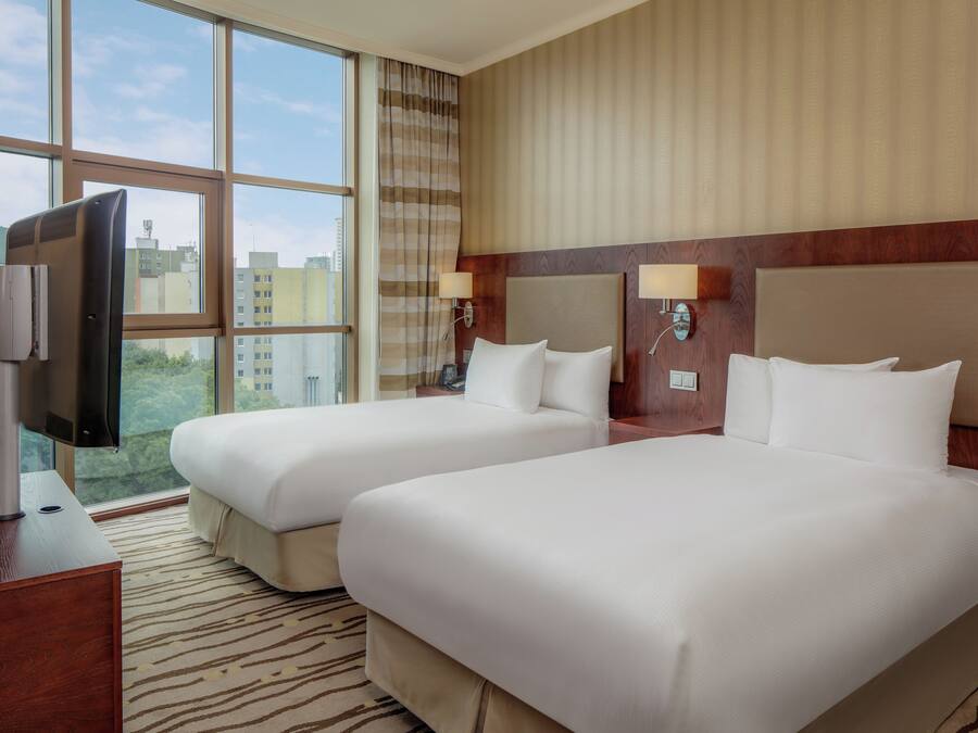 Quarto de hotel com duas camas, televisão e janelas amplas com vista para a cidade.