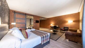 Una habitación de hotel moderna con una cama grande, una zona de estar con sofá y sillón, una mesita y un espejo.