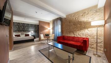 A modern hotel room with a wooden floor, a red sofa, a glass coffee table, a white bed with red pillows, a small table with chairs, and a stone wall.