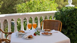 A table set with breakfast items, including croissants and juice, on a balcony with a white railing and greenery in the background.