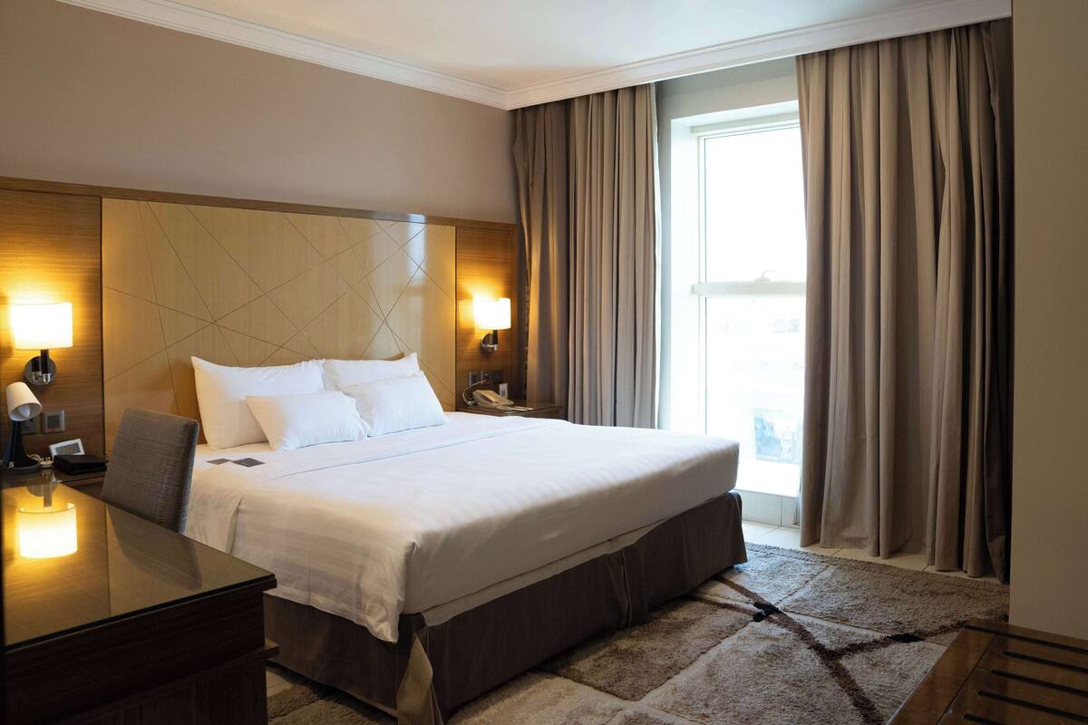 A hotel room with a large bed, two bedside lamps, a desk with a lamp, and a view of the outdoors through a window with curtains.