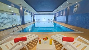 Une piscine intérieure à l’eau d’un bleu cristallin, entourée de chaises longues blanches et de serviettes rouges. Au centre, on trouve une table avec un panier de fruits et des boissons.