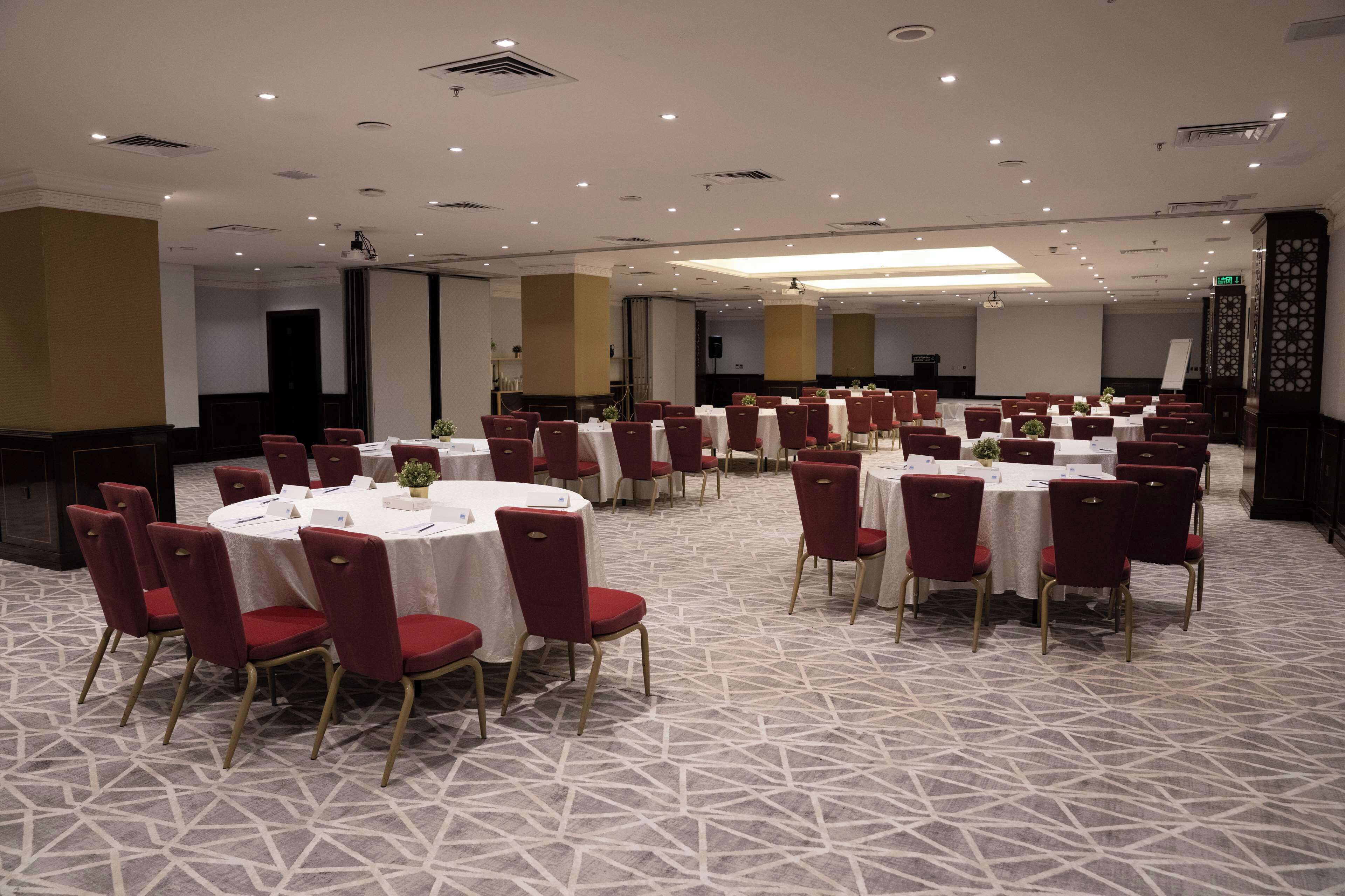 Un salón de conferencias con mesas redondas y sillas rojas, suelo alfombrado e iluminación empotrada.