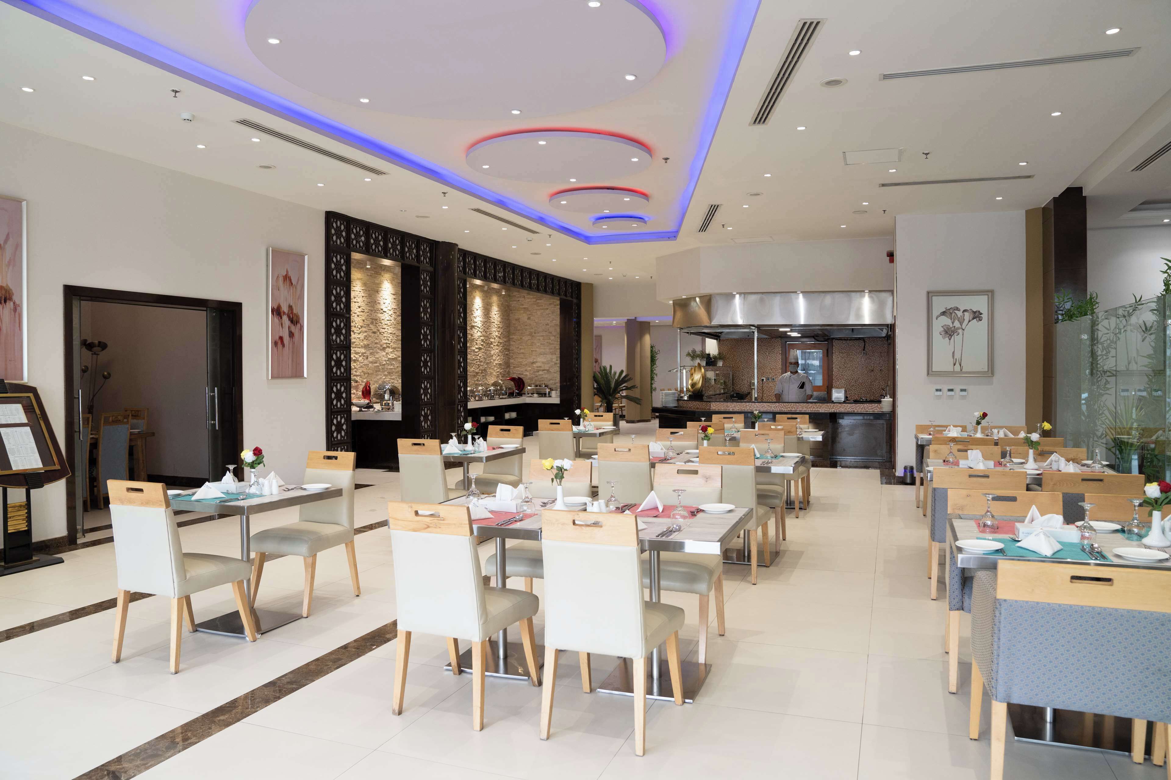 A modern dining area with multiple tables and chairs, a kitchen in the background, and decorative wall panels.