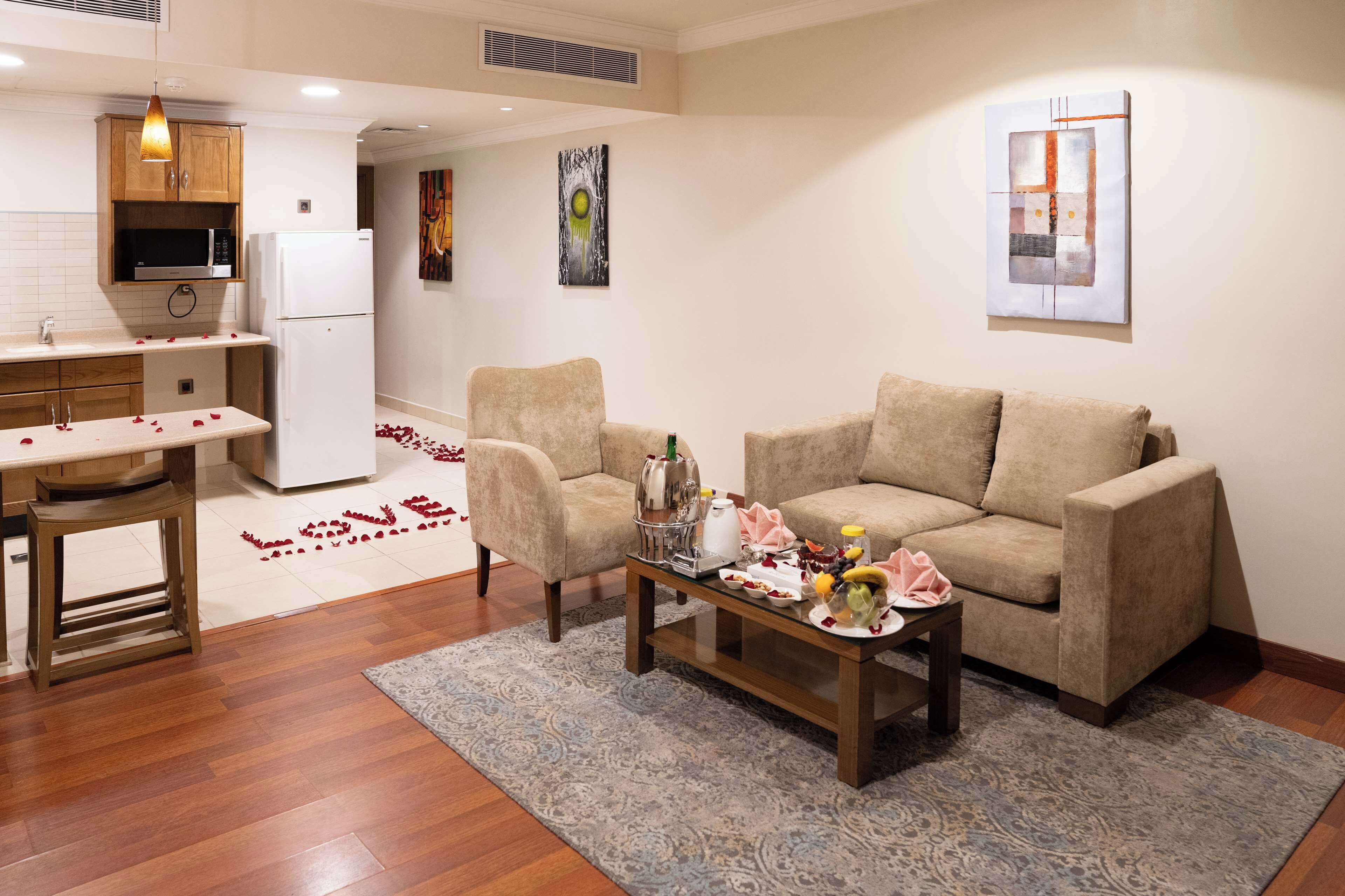 A modern living room with a beige sofa, a coffee table with a floral arrangement, and a kitchen area with wooden cabinets and a microwave.
