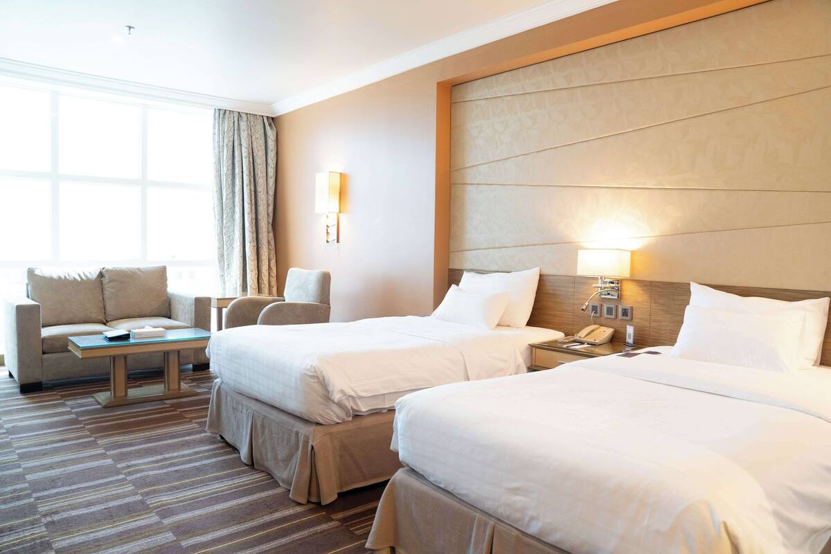 A hotel room with two beds, a sofa, a small table, and a nightstand with a lamp.