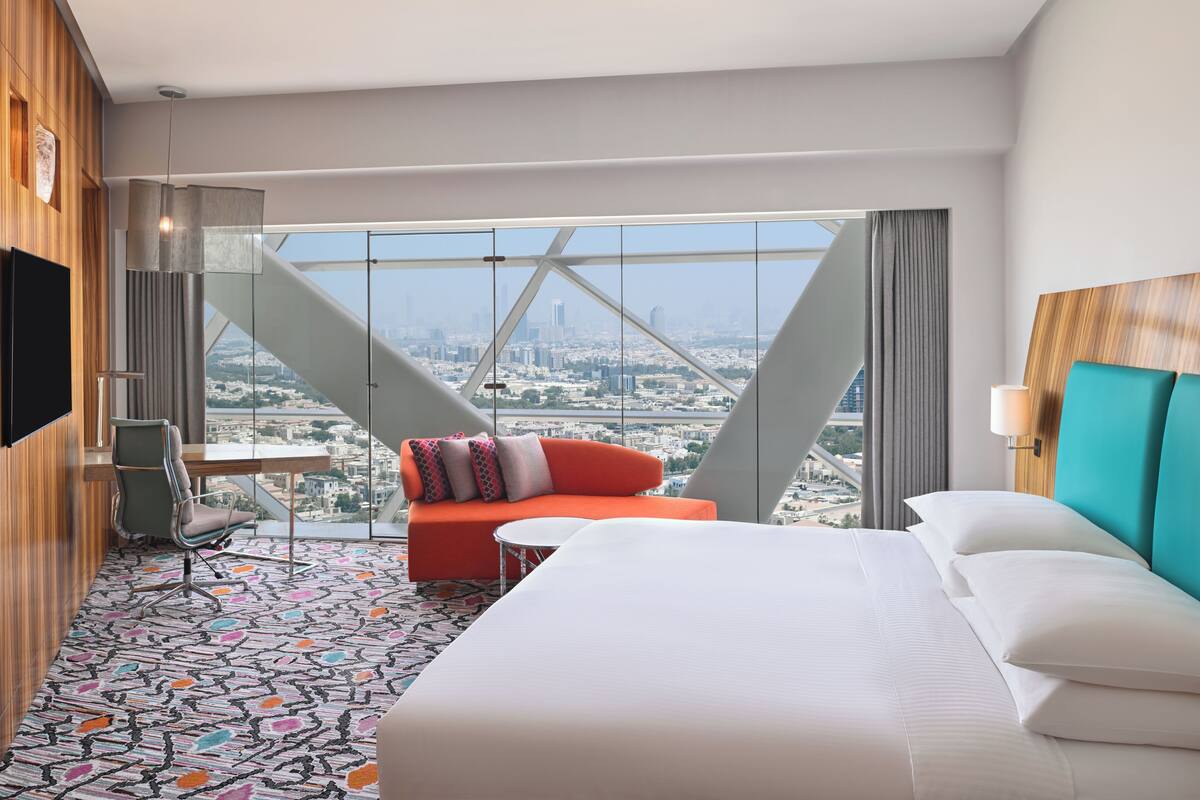 Ein Hotelzimmer mit einem großen Bett, einer Couch, einem Schreibtisch und einem Sessel. Durch große Fenster bietet das Zimmer Blick auf die Stadt.