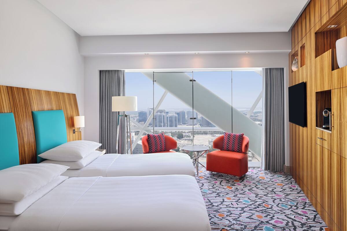 Ein Hotelzimmer mit zwei Betten, einem roten Sessel, einem kleinen Tisch und Blick auf die Stadt durch große Fenster.