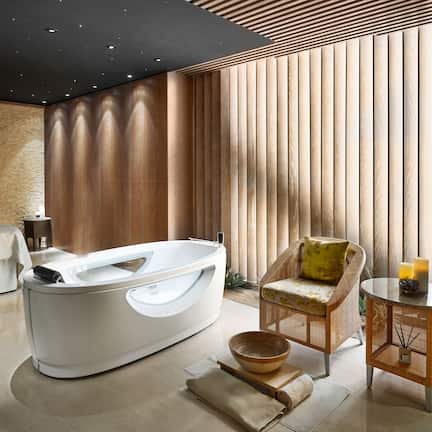 A modern spa room with a freestanding bathtub, a massage table, a wicker chair, and a small round table with candles.