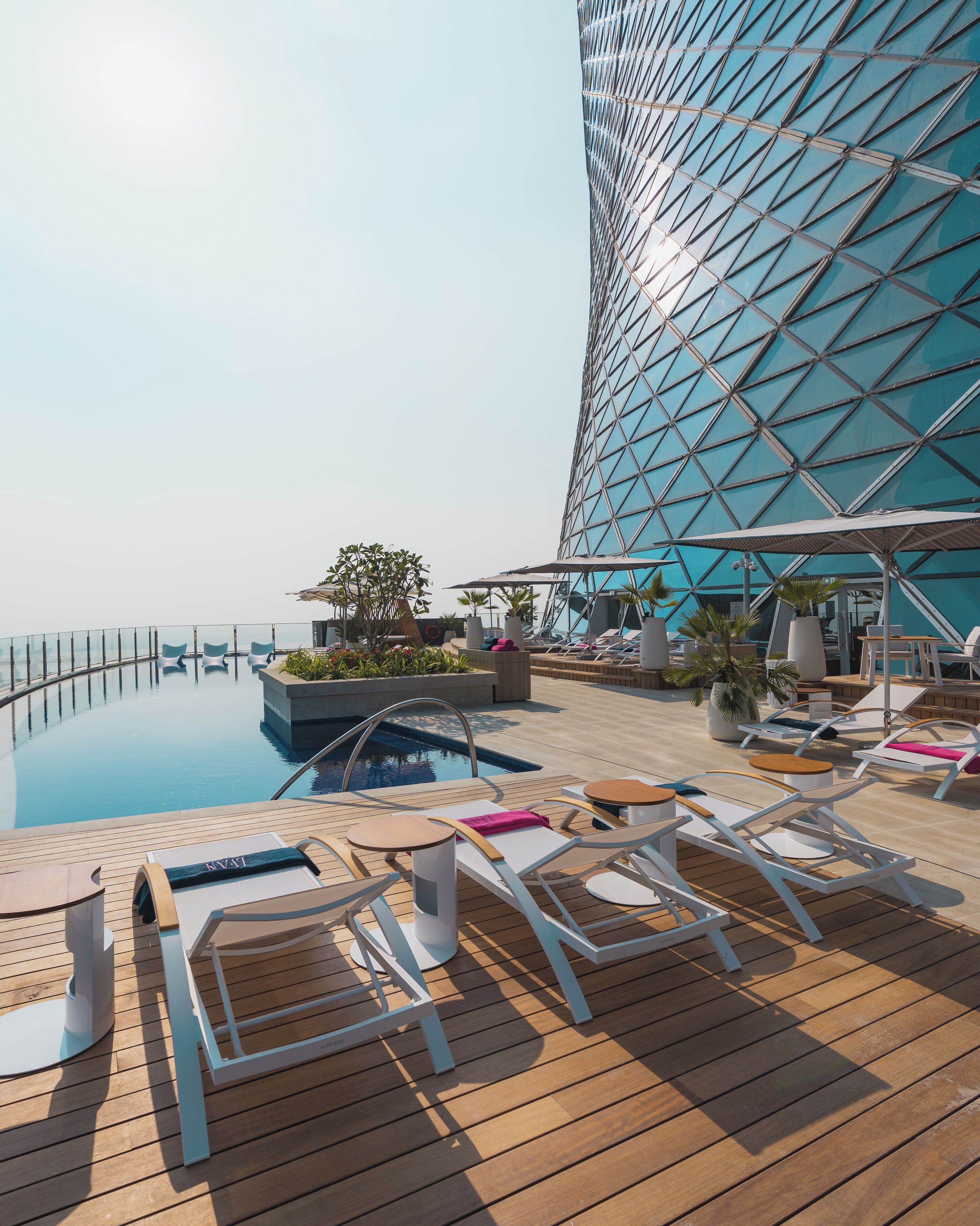 Un espace piscine aménagé sur le toit, avec des chaises longues, et un bâtiment moderne doté d’une façade en verre aux lignes géométriques.