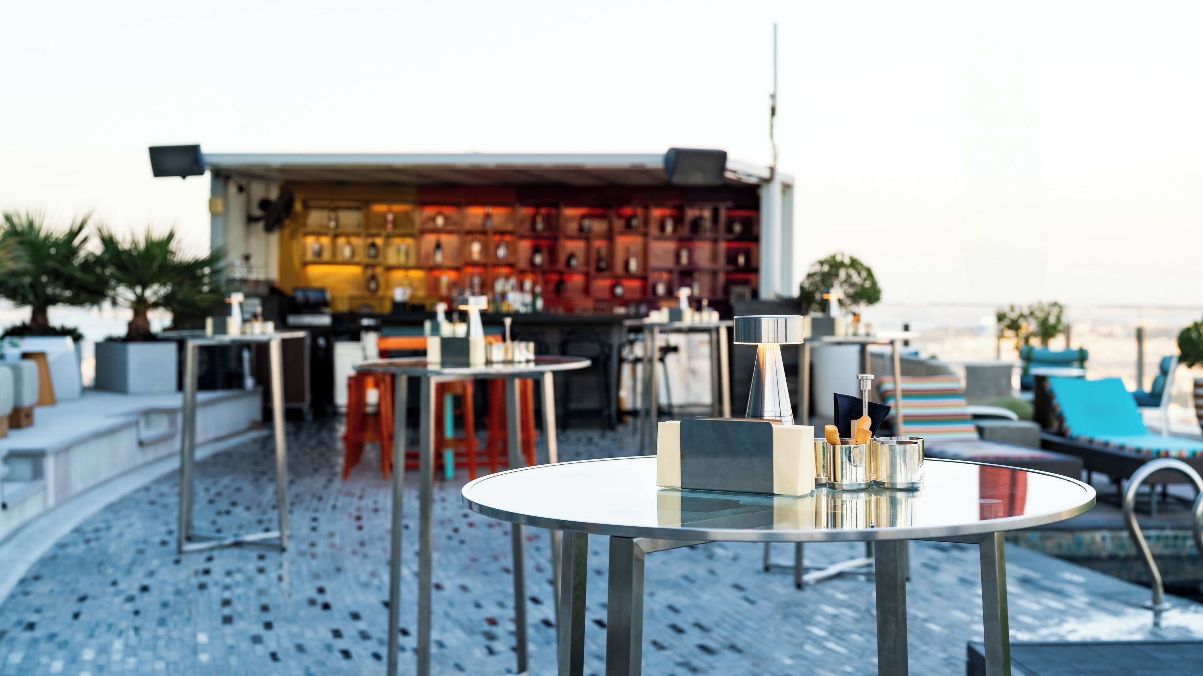 A rooftop bar with round tables, chairs, and a bar area with shelves of bottles.