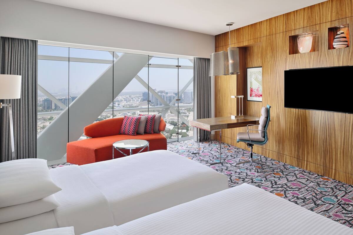 Ein Hotelzimmer mit einem großen Bett, einer Couch mit bunten Kissen, einem kleinen runden Tisch, einer Holzwand als Blickfang, einem an der Wand befestigten Fernseher und einem Blick auf die Stadt durch große Fenster.