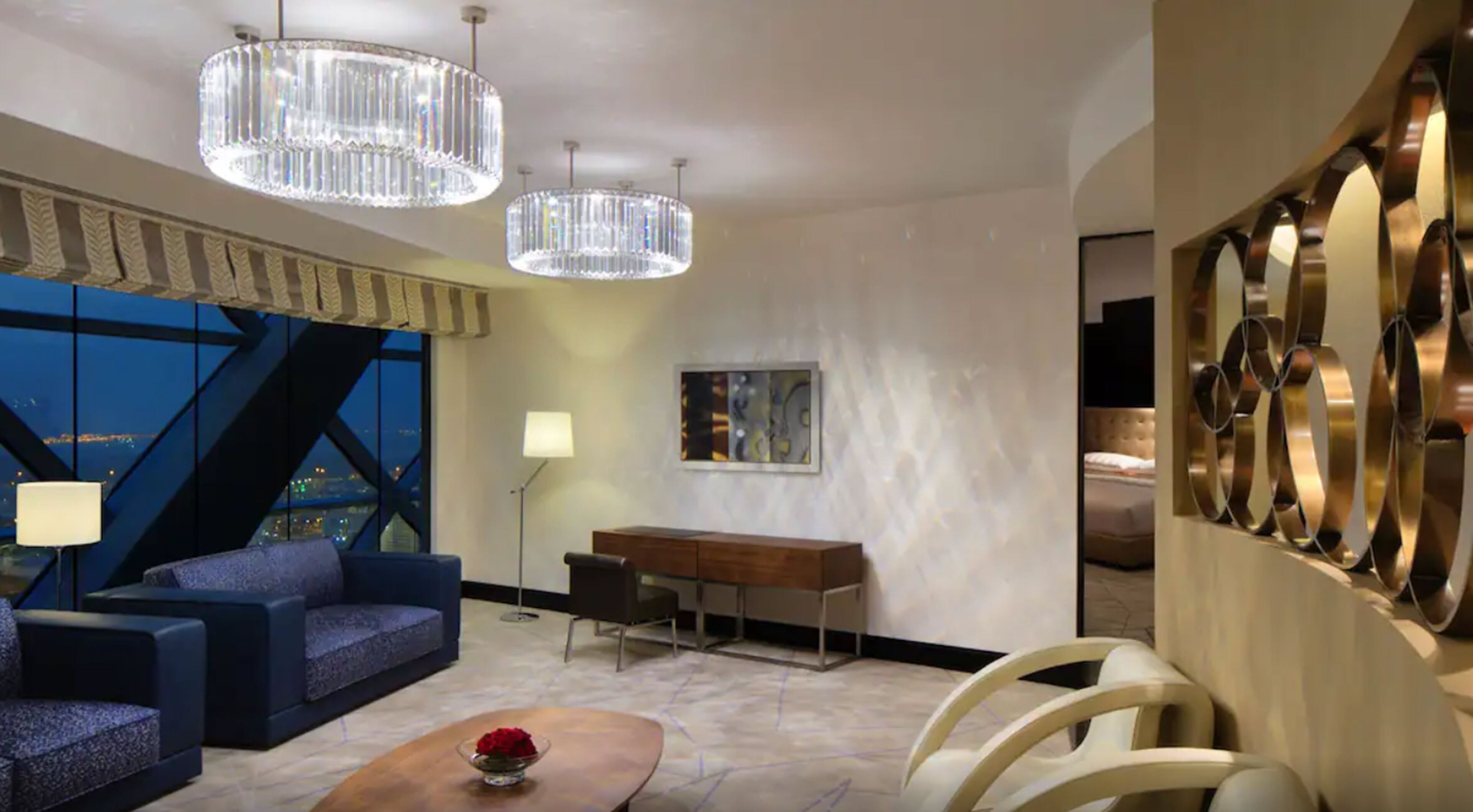 A modern hotel room with a large window, a blue sofa, a wooden desk, and a decorative wall piece.