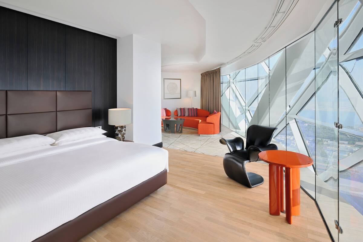 Ein modernes Hotelzimmer mit einem großen Bett, einer Glaswand mit Blick auf die Stadt, einer Sitzecke mit orangefarbenen Möbeln und einem dunklen Kopfteil.