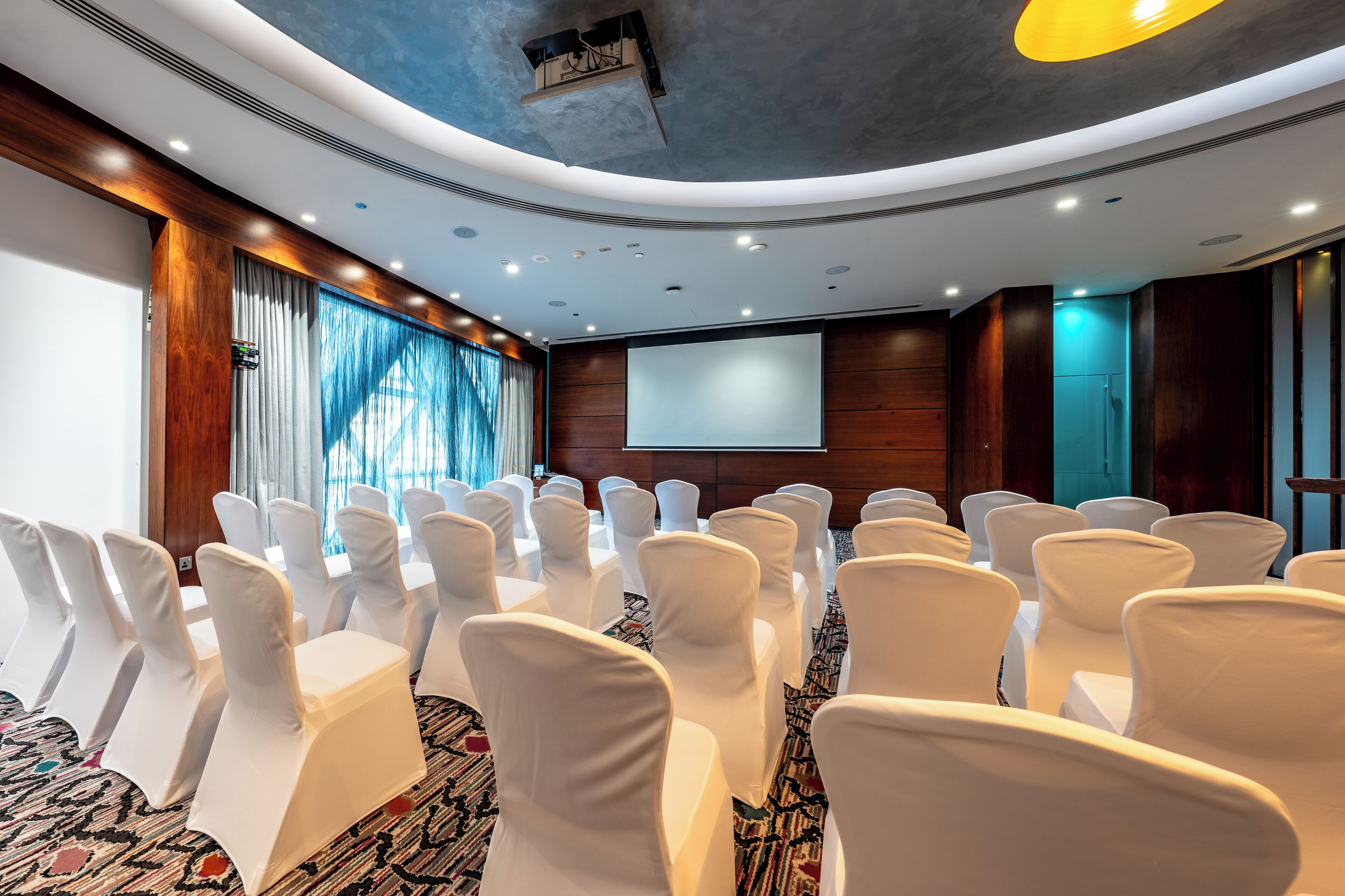A conference room with rows of white chairs, a large screen, and a wooden panel wall.