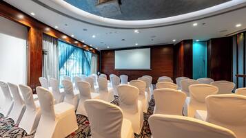 A conference room with rows of white chairs, a large screen, and a wooden panel wall.