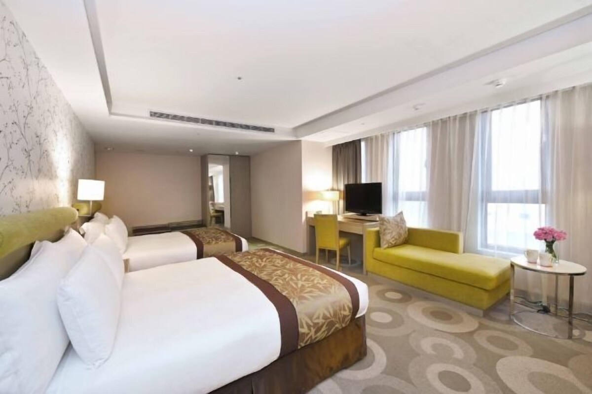 Une chambre d’hôtel avec deux lits, un canapé jaune, une télévision et une petite table sur laquelle est posé un vase de fleurs.