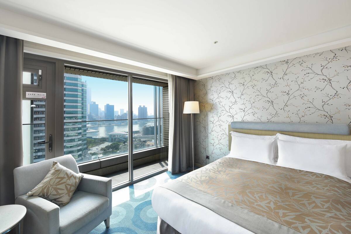 Een hotelkamer met een groot bed, een bank en uitzicht op de stad door middel van kamerhoge ramen.