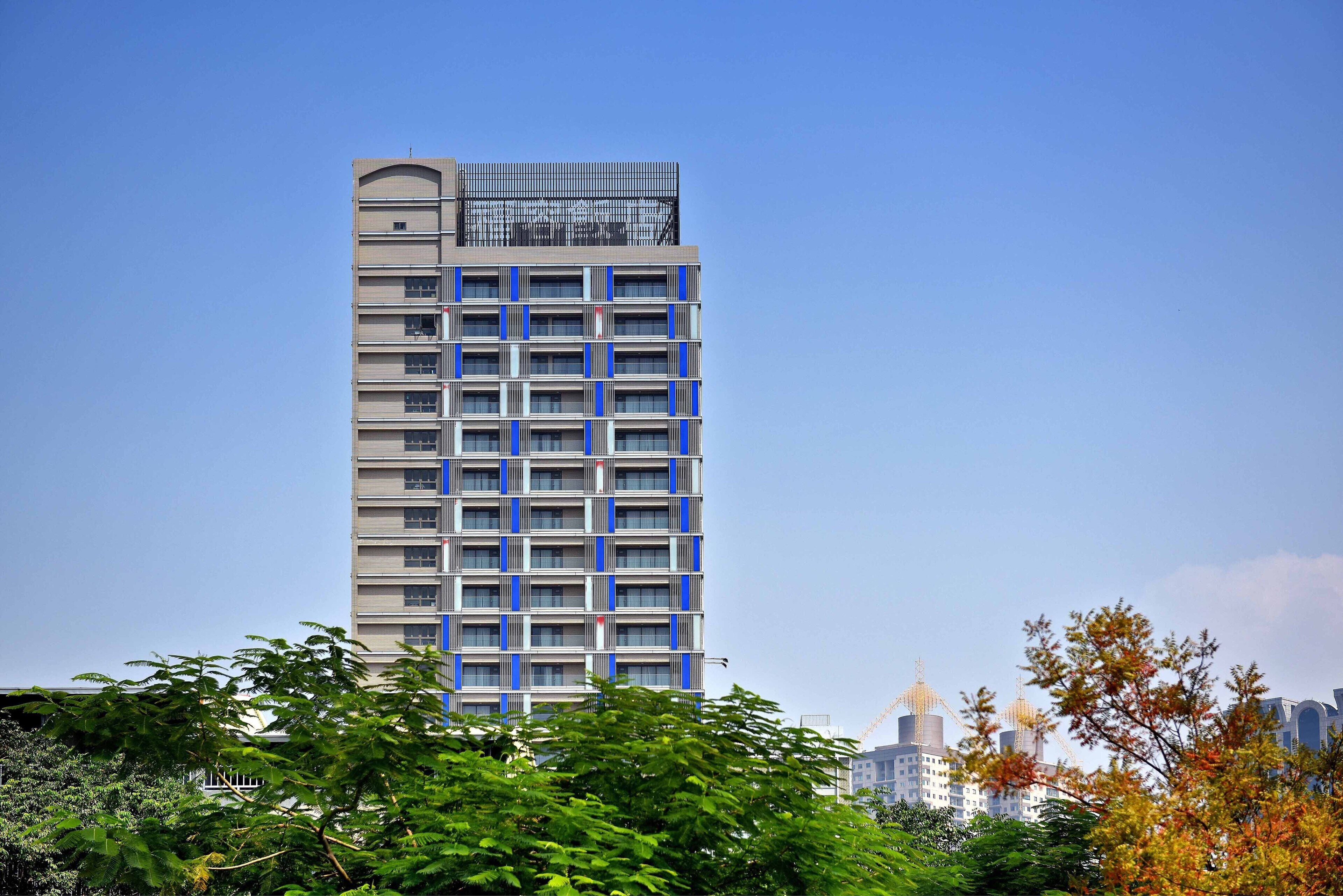A tall, modern building with a grid of windows and balconies, surrounded by trees and other buildings.