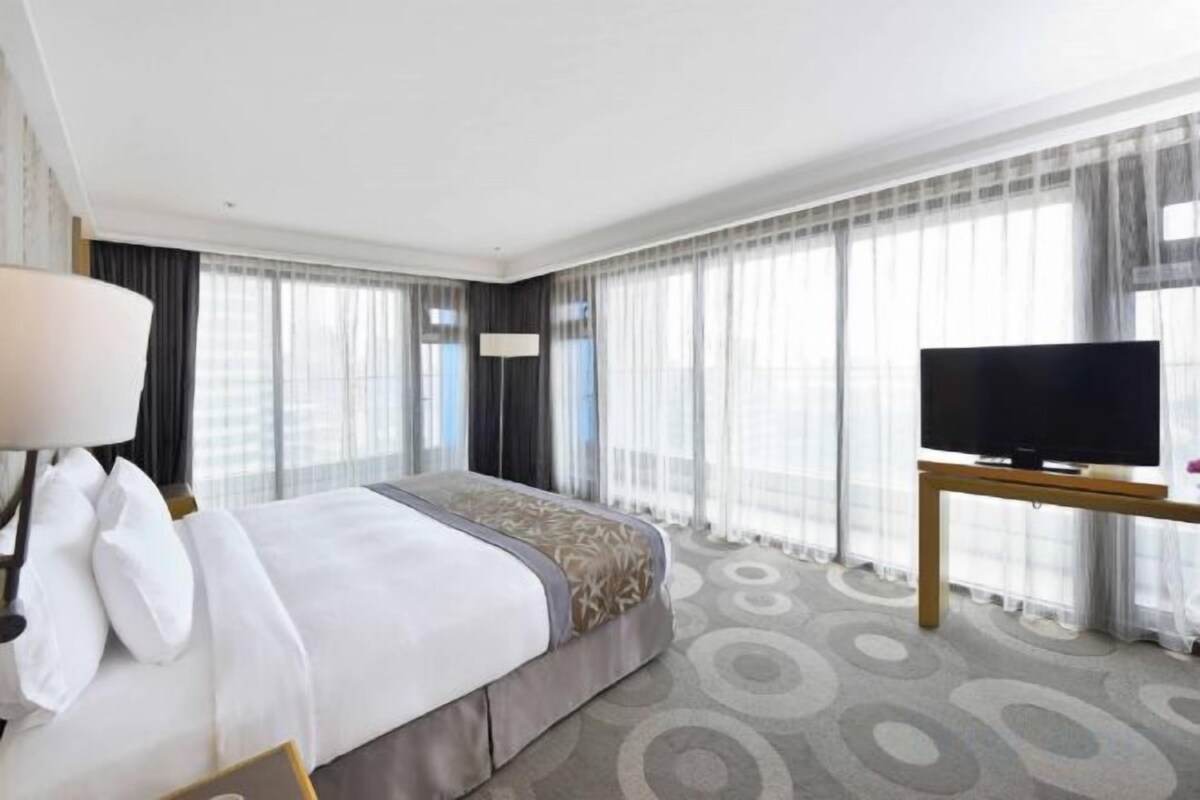 Ein Hotelzimmer mit einem Bett, einem Fernseher auf einem Tisch und großen Fenstern mit Vorhängen.