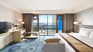 Ein Hotelzimmer mit einem großen Bett, einem Schreibtisch mit Fernseher, einem Sessel, einem kleinen Tisch mit einer Vase und Blick auf die Stadt durch große Fenster.