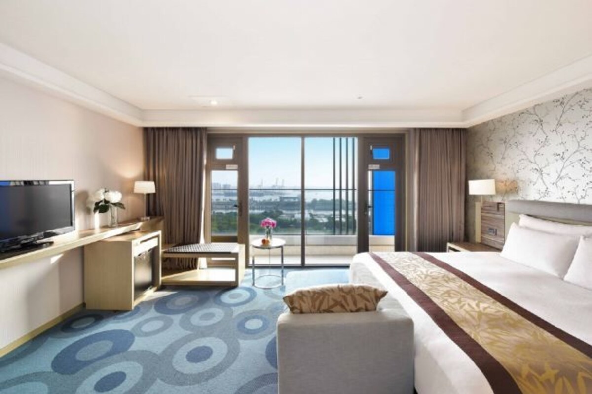 Une chambre d’hôtel avec un grand lit, un bureau avec une télévision, une petite table avec une lampe, et une vue sur le paysage urbain à travers de grandes fenêtres.