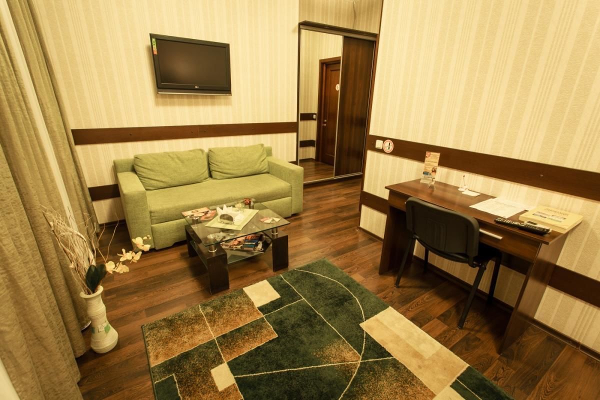 A compact hotel room with a green sofa, a small coffee table, a desk with a chair, a flat-screen TV mounted on the wall, and a mirror above the door.