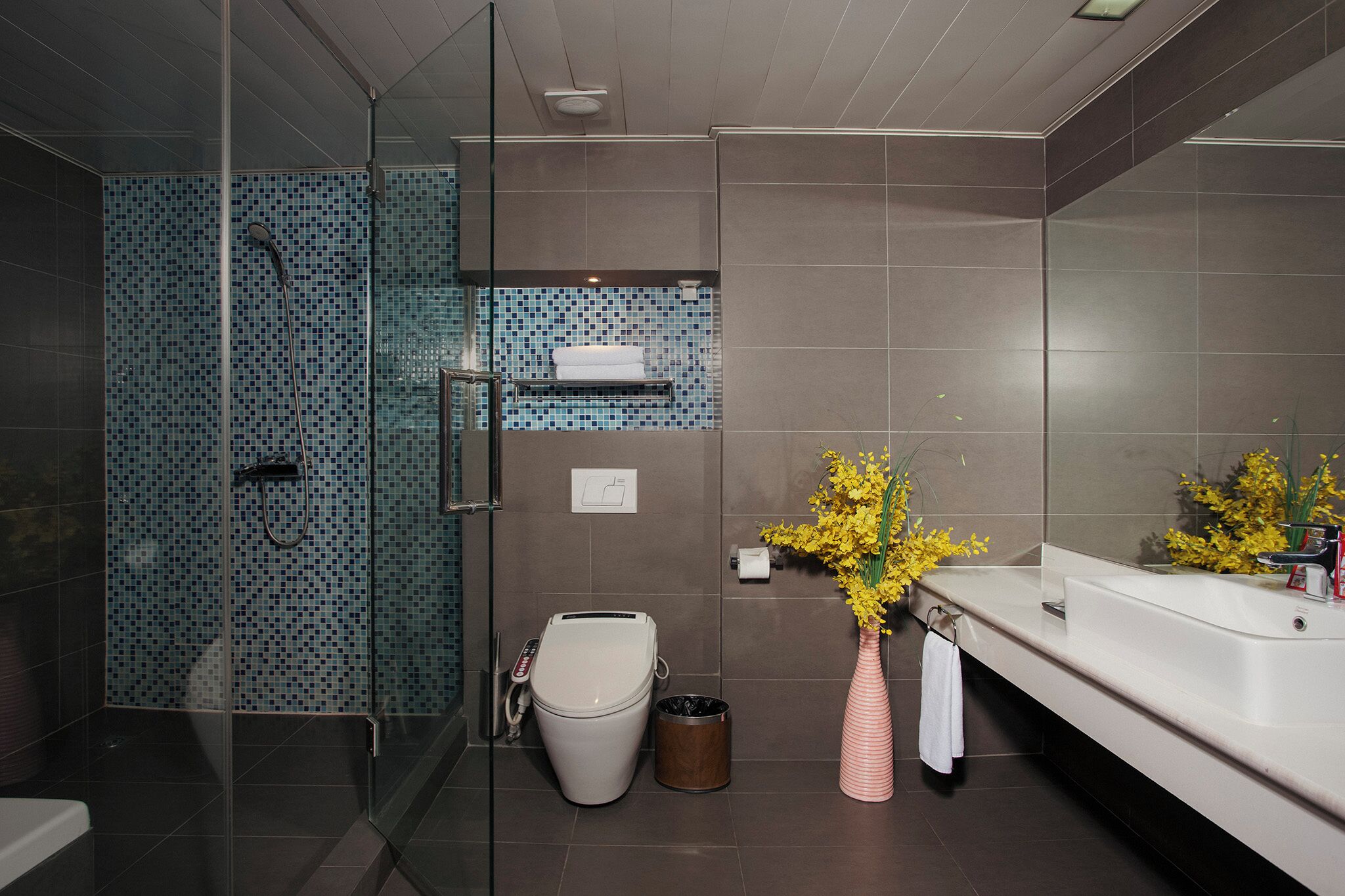 A modern bathroom with a glass shower enclosure, a toilet, a sink with a mirror, and a vase with yellow flowers.