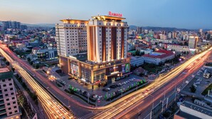 Un hotel moderno de muchas plantas con un llamativo letrero de 'RAMADA', rodeado de un entorno urbano concurrido donde se ve movimiento de vehículos y otros edificios.