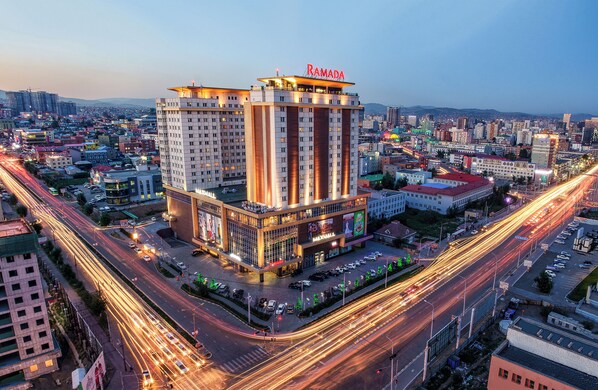 A high-rise hotel with a prominent 'RAMADA' sign, surrounded by a busy urban environment with visible traffic and other buildings. - Ramada by Wyndham Ulaanbaatar Citycenter (Ulaanbaatar)