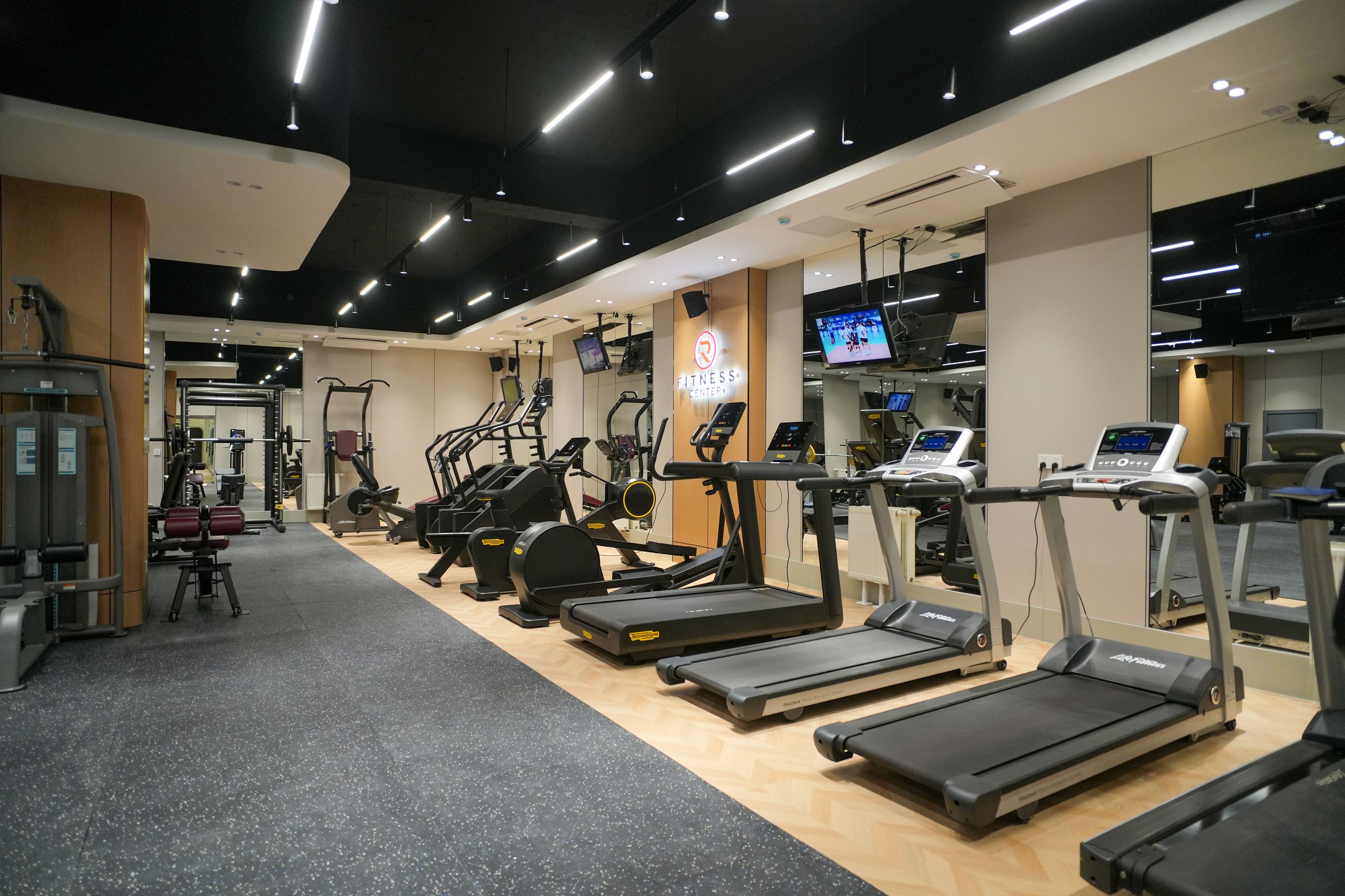 A modern gym with various cardio machines, weightlifting equipment, and a digital display.