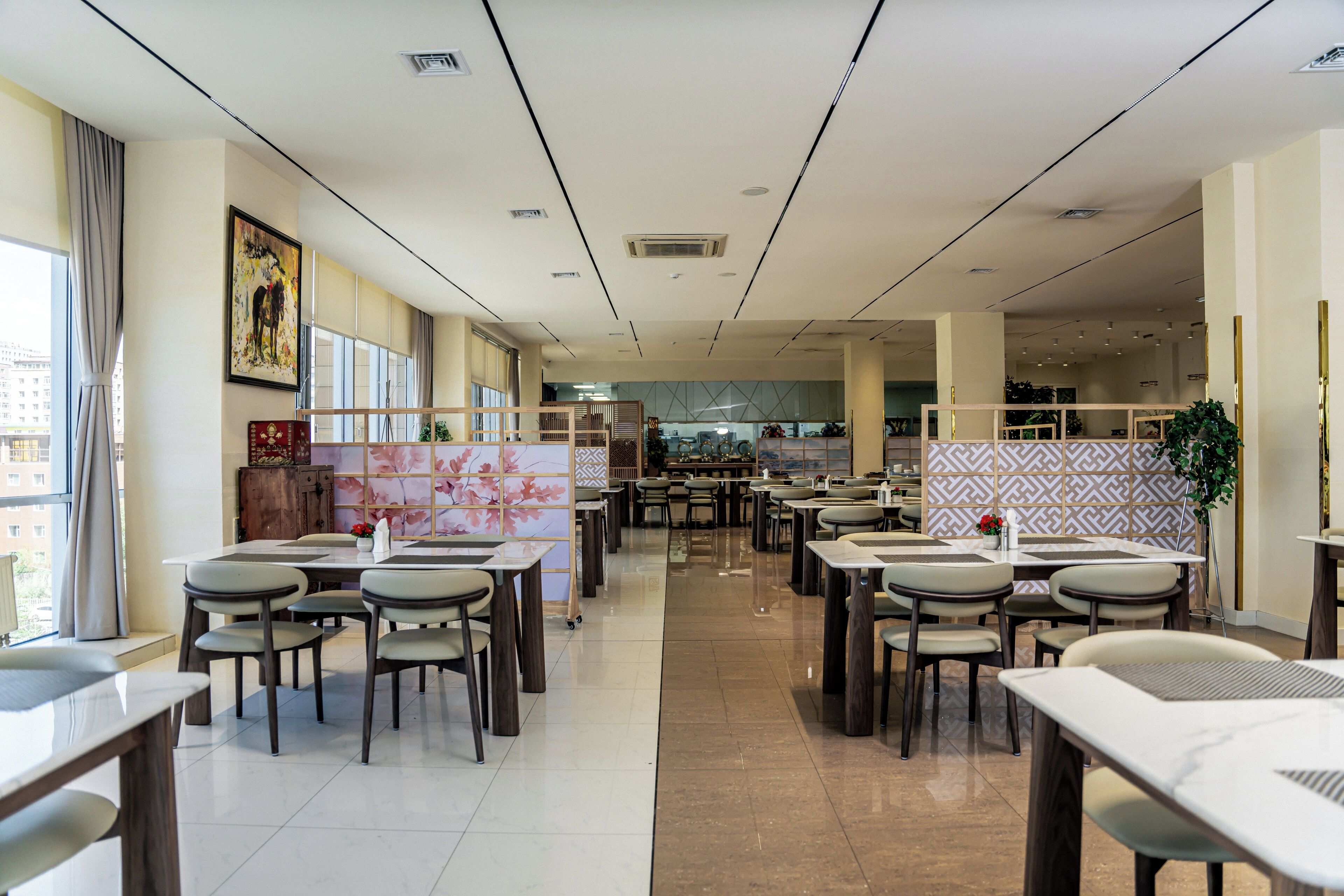 A spacious restaurant with multiple dining tables and chairs, a large window on the left, and a patterned wall in the background.