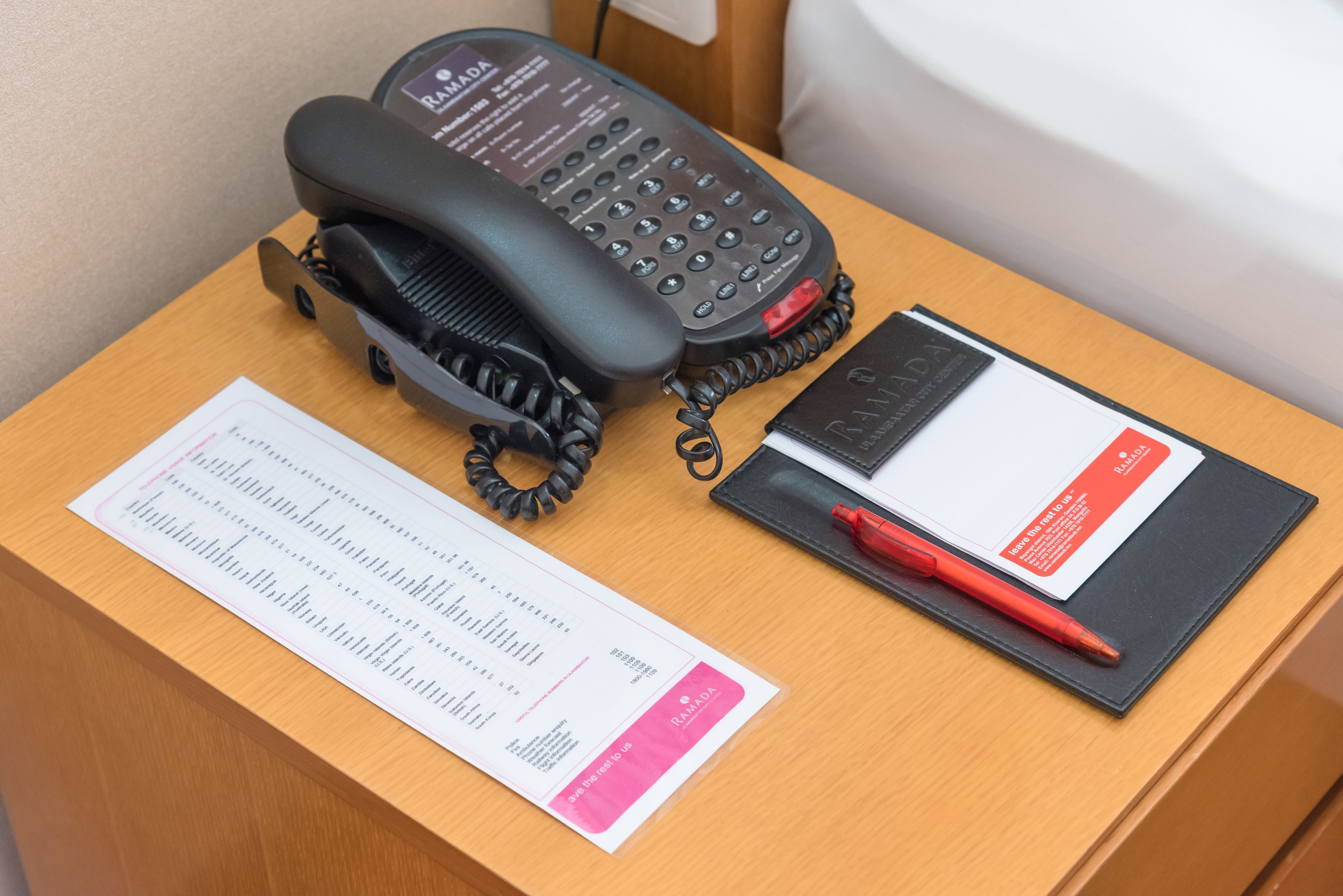 A wooden desk with a black telephone, a pink and white document, and a red pen.
