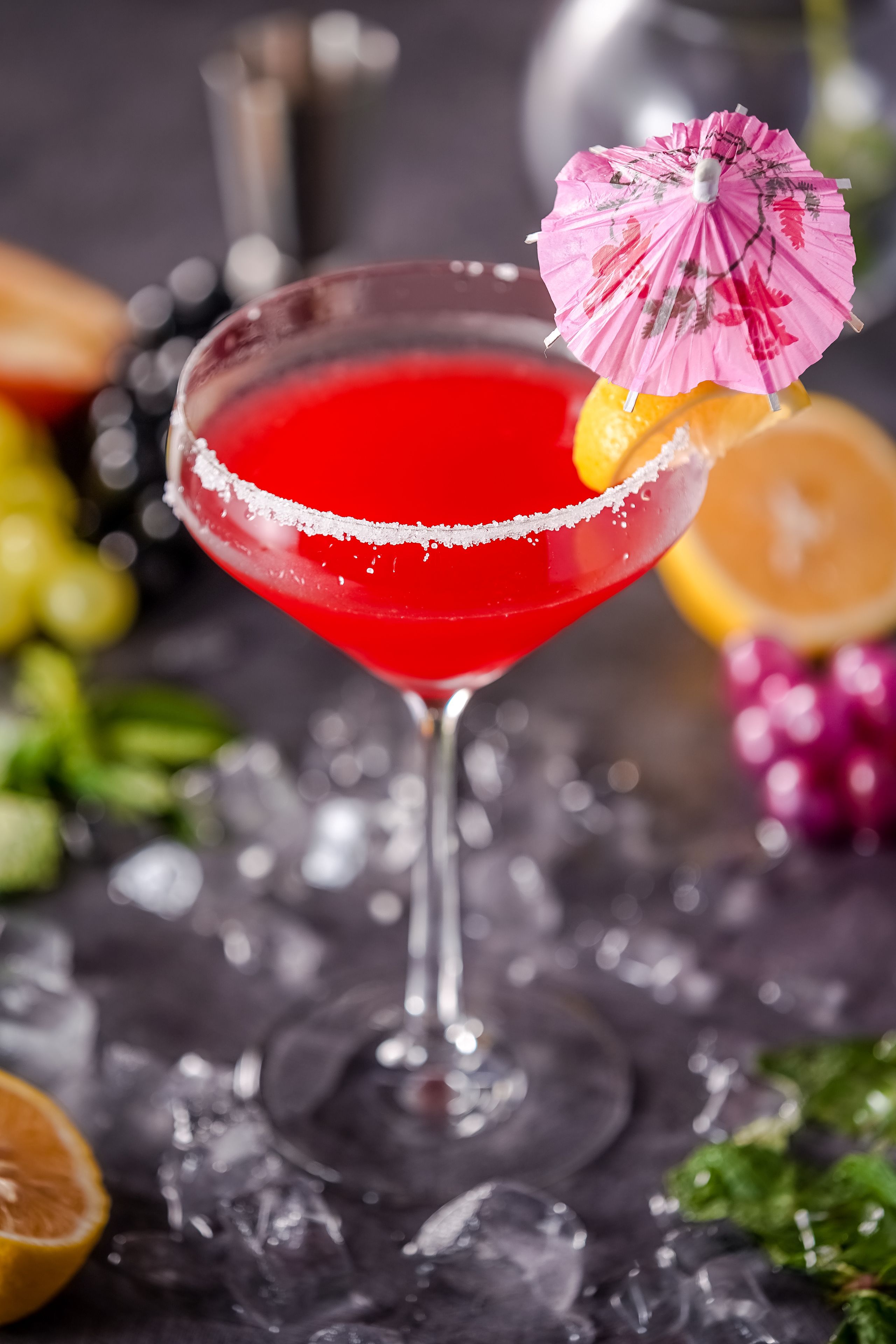 A cocktail with a pink umbrella garnish on a glass with a salted rim, surrounded by ice cubes and citrus fruits.