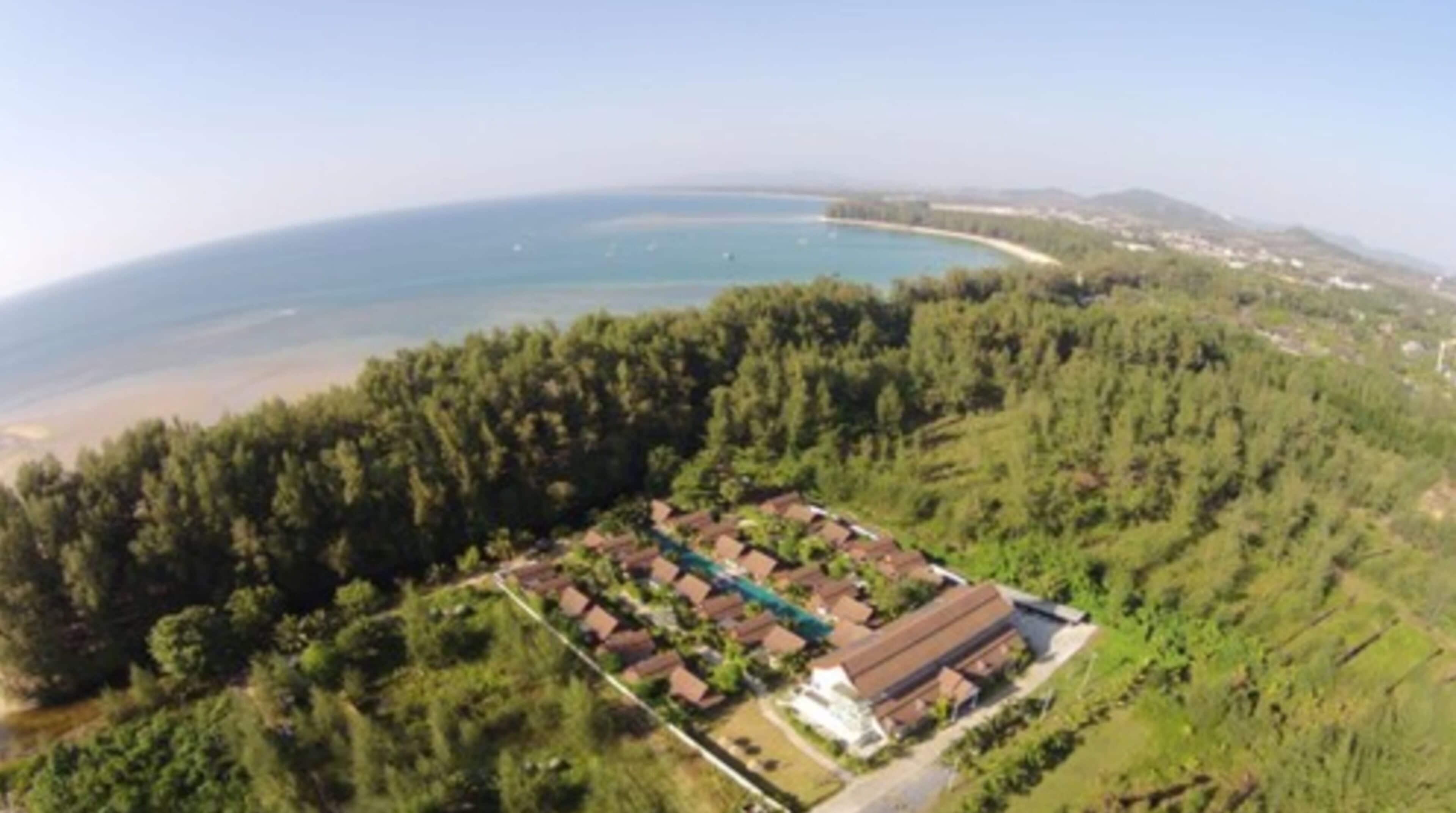 An aerial view of a resort with a large building surrounded by dense forest, overlooking a beach and the ocean.