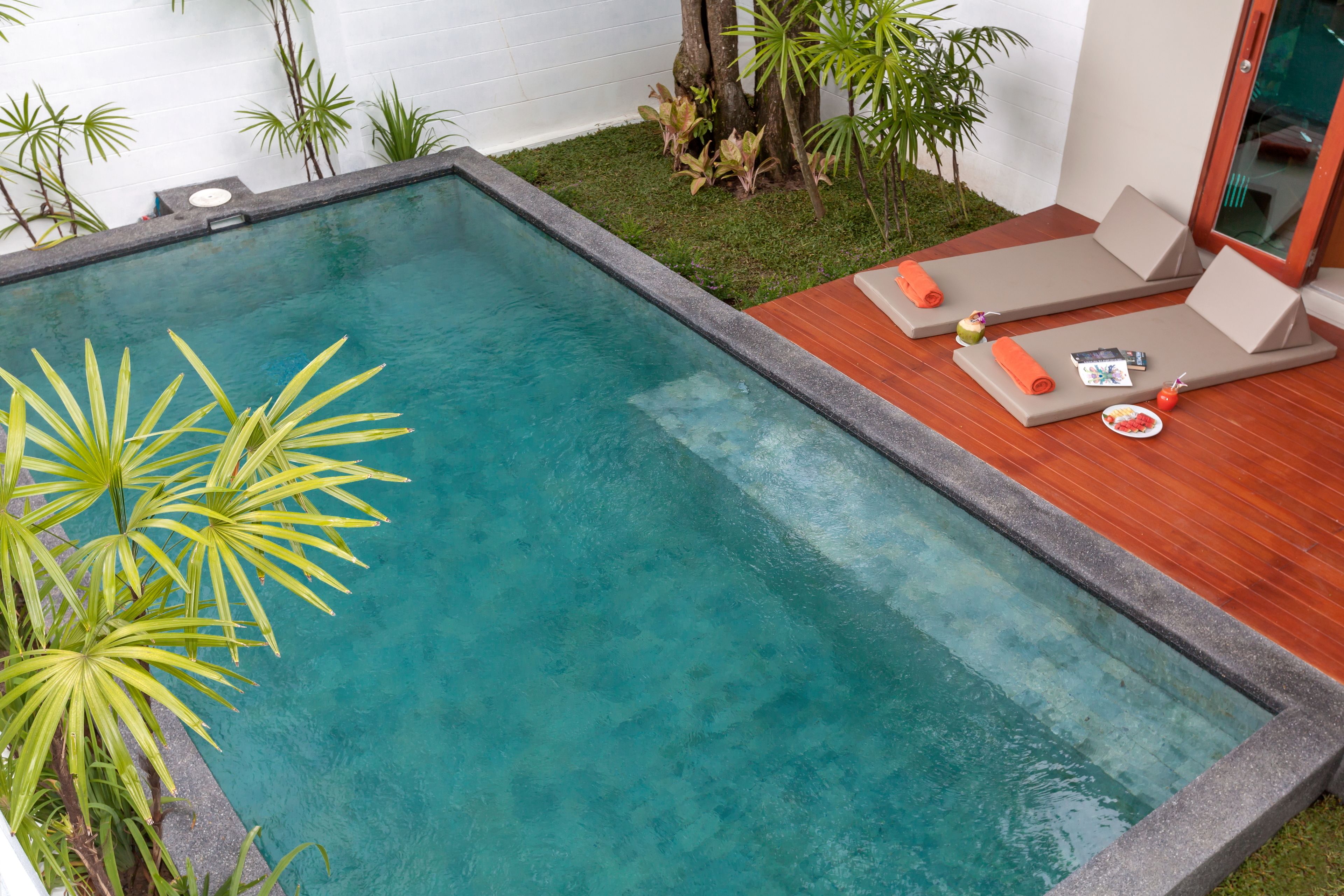 A rectangular swimming pool with a stone edge, surrounded by a wooden deck and a small grassy area with plants.