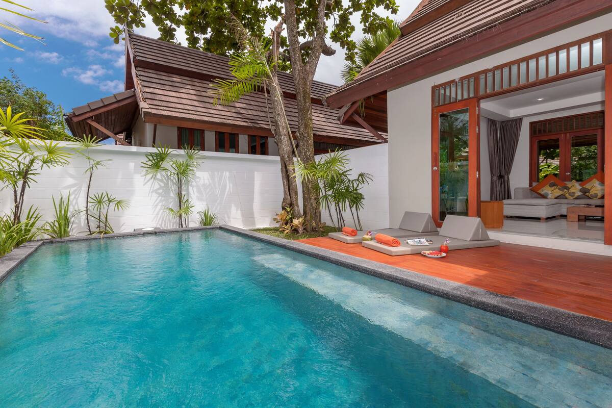 A swimming pool with a wooden deck and lounge chairs, surrounded by a white wall and tropical plants.